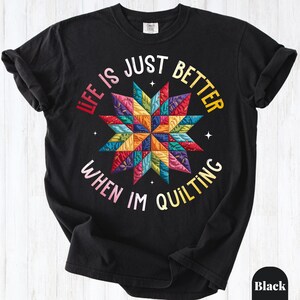 May include: A black short-sleeve t-shirt featuring a colorful, multi-patterned star design in the center. The text "LIFE IS JUST BETTER WHEN IM QUILTING" is printed in a circular arrangement around the star in shades of red, yellow, and white.