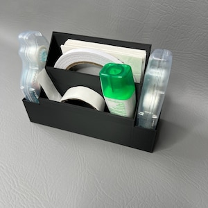 May include: A black organizer with three compartments holds various office supplies, including rolls of tape, a bottle of glue, and a stack of white paper.