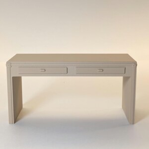 May include: A light beige desk with two drawers and a rectangular top. The desk has a simple, modern design with decorative trim around the edges and drawer fronts. The desk is made of a solid material and has a neutral colour.