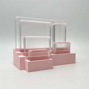 May include: A pink desk organizer with multiple compartments. Clear acrylic rectangles of varying sizes are arranged in the organizer. The organizer has a stepped design and is made of solid pink material.