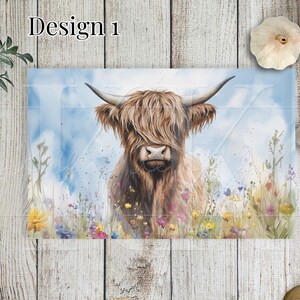 May include: A Highland cow with long brown fur stands in a field of wildflowers. The cow is looking directly at the viewer with a gentle expression. The background is a soft blue watercolour wash.