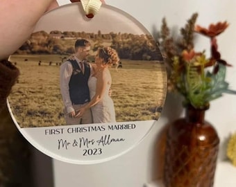 May include: A round, clear glass ornament with a photo of a couple in a field. The ornament has the text "First Christmas Married Mr. & Mrs. Allman 2023" printed on it.