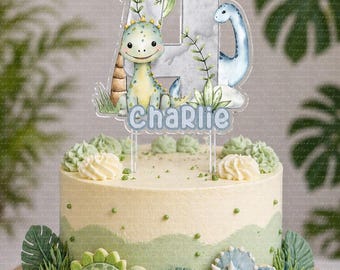 Könnte beinhalten: Ein dekorierter Kuchen mit Dinosaurier-Thema. Der Kuchen hat ein hellgrünes und weißes Frosting mit grünen Akzenten. Ein klarer Aufsatz mit der Zahl 4, Dinosauriern und dem Namen "Charlie" befindet sich oben. Zusätzliche Dinosaurierfiguren und Blätter schmücken die Basis.