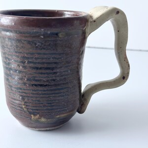 May include: A handmade ceramic mug with a textured, dark brown exterior and a cream-colored handle. The mug features horizontal lines and a rustic aesthetic, perfect for enjoying coffee or tea. The mug's dimensions are approximately 4 inches tall.