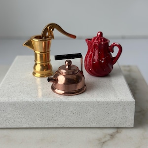 May include: A collection of miniature kitchenware items, including a gold-colored espresso maker, a copper-colored kettle with a black handle, and a red ceramic pitcher with white speckles. The items are arranged on a white surface.