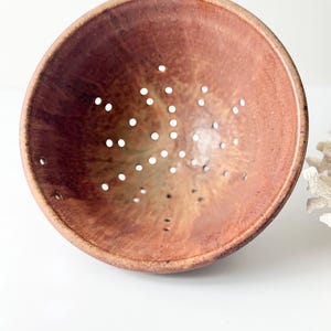 May include: A round ceramic colander featuring a mottled terracotta and brown glaze with a smooth finish. The bowl-shaped vessel has a series of small, evenly spaced drainage holes distributed across its interior surface and base.