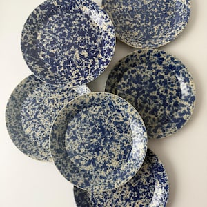 Può includere: Un set di sei piatti rotondi in ceramica con un motivo blu e crema maculato. I piatti sono disposti in modo leggermente sovrapposto, mostrando il loro design. I piatti hanno un'estetica classica e vintage.