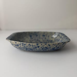 May include: A speckled, square-shaped ceramic bowl. The bowl has a light beige base with dark blue speckles. The rim is slightly raised, and the bowl is empty. The bowl is likely for serving food.
