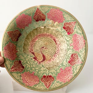 May include: A round brass decorative plate featuring a central peacock motif with an expanded tail. The surface is decorated with a light green enamel background and intricate floral patterns in deep pink and gold, creating a detailed design.