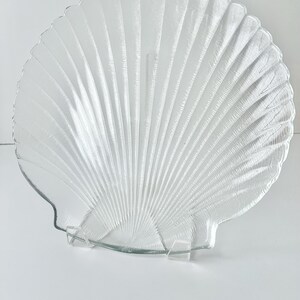 May include: A clear glass seashell-shaped serving dish. The dish has a scalloped edge and a textured, ribbed surface, resembling the natural lines of a seashell. It is displayed on a clear stand, perfect for serving appetizers or displaying decorative items.