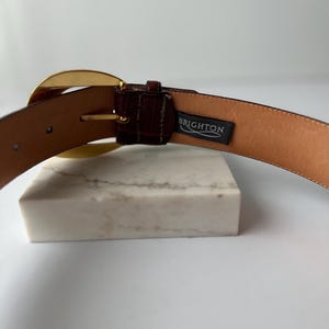 Puede incluir: Un cintur&oacute;n de cuero marr&oacute;n con una hebilla dorada. El cintur&oacute;n tiene un acabado texturizado y una etiqueta que dice "BRIGHTON". El cintur&oacute;n se muestra sobre una superficie blanca.