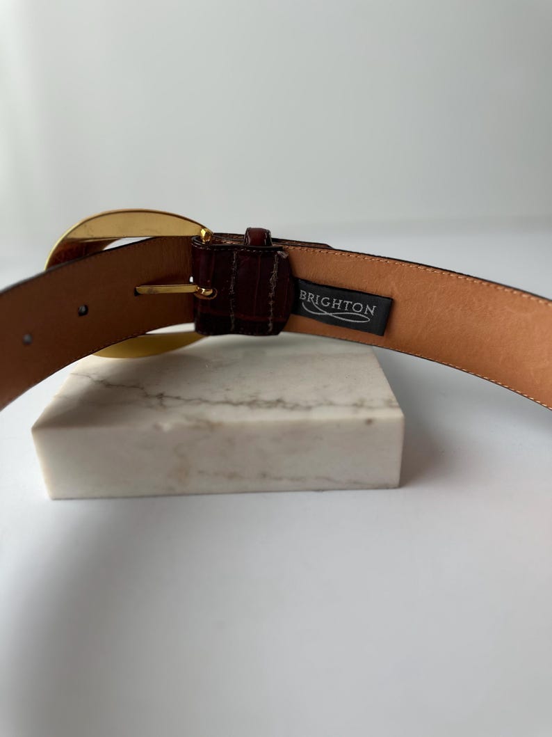 Puede incluir: Un cintur&oacute;n de cuero marr&oacute;n con una hebilla dorada. El cintur&oacute;n tiene un acabado texturizado y una etiqueta que dice "BRIGHTON". El cintur&oacute;n se muestra sobre una superficie blanca.