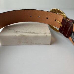 Puede incluir: Un cintur&oacute;n de cuero marr&oacute;n con una hebilla dorada. El cintur&oacute;n presenta una secci&oacute;n texturizada, marr&oacute;n oscuro cerca de la hebilla y un interior de cuero marr&oacute;n claro. El cintur&oacute;n se muestra sobre una superficie blanca.