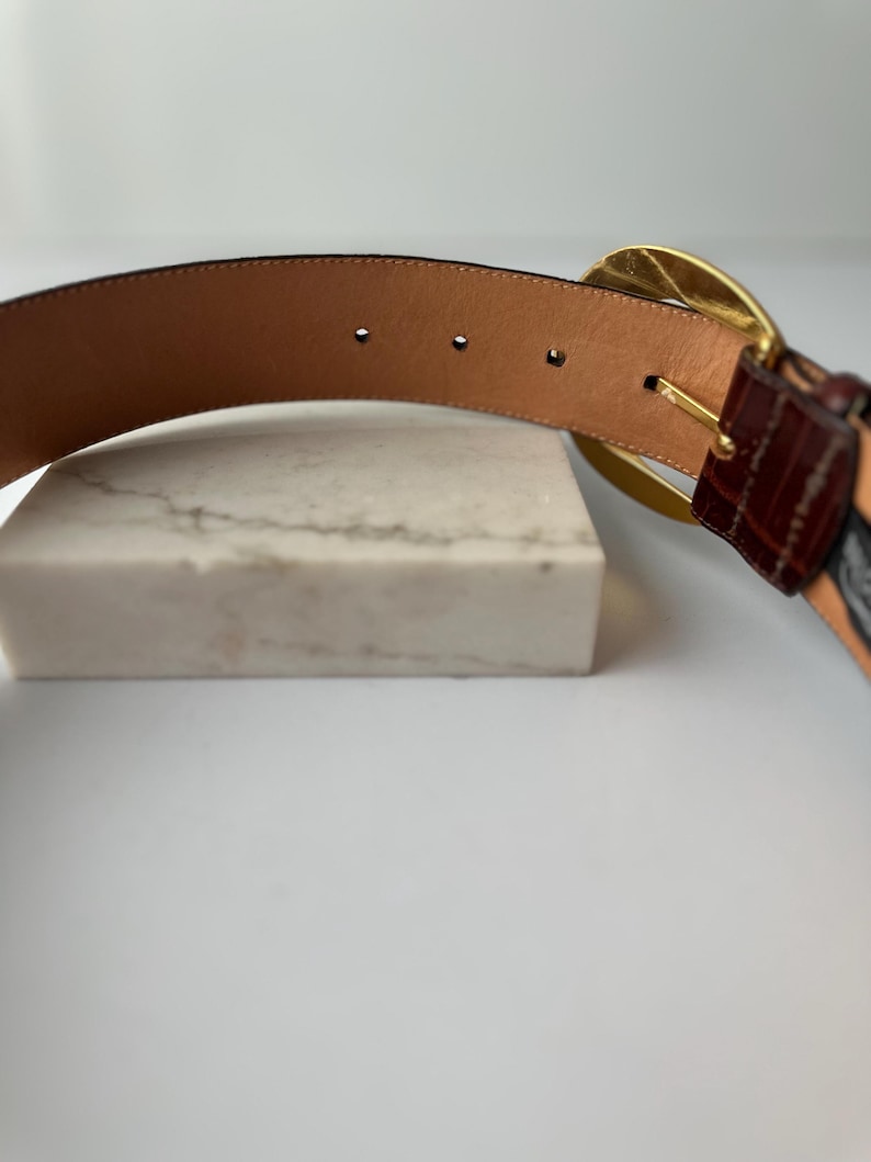 Puede incluir: Un cintur&oacute;n de cuero marr&oacute;n con una hebilla dorada. El cintur&oacute;n presenta una secci&oacute;n texturizada, marr&oacute;n oscuro cerca de la hebilla y un interior de cuero marr&oacute;n claro. El cintur&oacute;n se muestra sobre una superficie blanca.