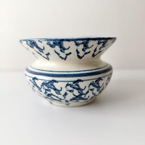 May include: A small, round ceramic bowl with a wide, flared rim. The off-white base features a hand-painted blue pattern consisting of repeating abstract, jagged marks and a solid horizontal blue line encircling the center of the vessel.