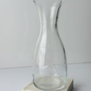 Op de afbeelding: Een heldere glazen karaf met een brede basis en een smalle hals, op een witte marmeren blok. De karaf heeft een afgeronde body en een uitlopende bovenkant. Het woord "MILK" is zichtbaar op de karaf.
