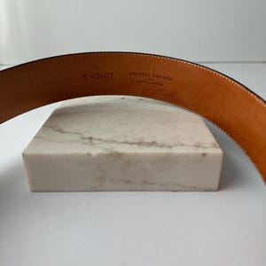 Puede incluir: Primer plano de un cintur&oacute;n de cuero marr&oacute;n. El texto "EMBOSSED CALFSKIN ON SADDLE LEATHER" y el n&uacute;mero "S 43407" est&aacute;n estampados en el interior. El cintur&oacute;n est&aacute; curvado y parece nuevo.