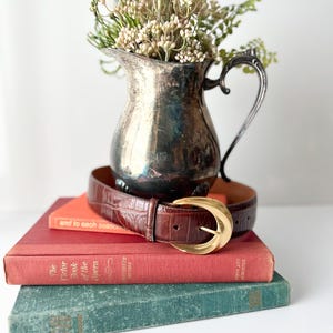 Puede incluir: Un cintur&oacute;n de cuero marr&oacute;n con hebilla dorada descansa sobre una pila de libros antiguos. Una jarra plateada llena de flores secas se encuentra encima de los libros. Los libros son en tonos rojos, naranjas y verdes.