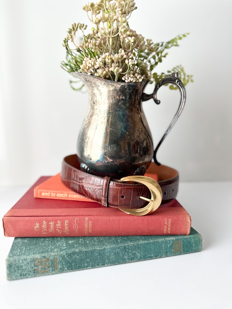 Puede incluir: Un cintur&oacute;n de cuero marr&oacute;n con hebilla dorada descansa sobre una pila de libros antiguos. Una jarra plateada llena de flores secas se encuentra encima de los libros. Los libros son en tonos rojos, naranjas y verdes.