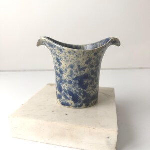 May include: A blue and cream-colored ceramic vase with a flared top. The vase has a speckled pattern of dark blue spots on a cream background. The vase is sitting on a white rectangular block.