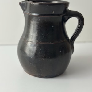 May include: A small, dark brown ceramic pitcher with a rounded body and a curved handle. The pitcher has a spout for pouring and a horizontal line detail around the middle. The surface has a glossy finish.