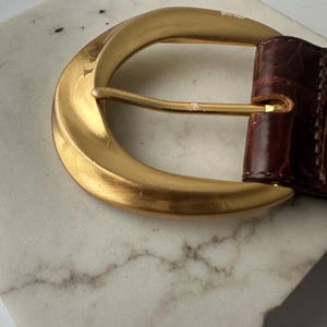 Puede incluir: Primer plano de un cintur&oacute;n de cuero marr&oacute;n con una hebilla grande de color dorado. La hebilla tiene forma de media luna curvada y un dise&ntilde;o simple y elegante. El cintur&oacute;n de cuero parece tener textura.