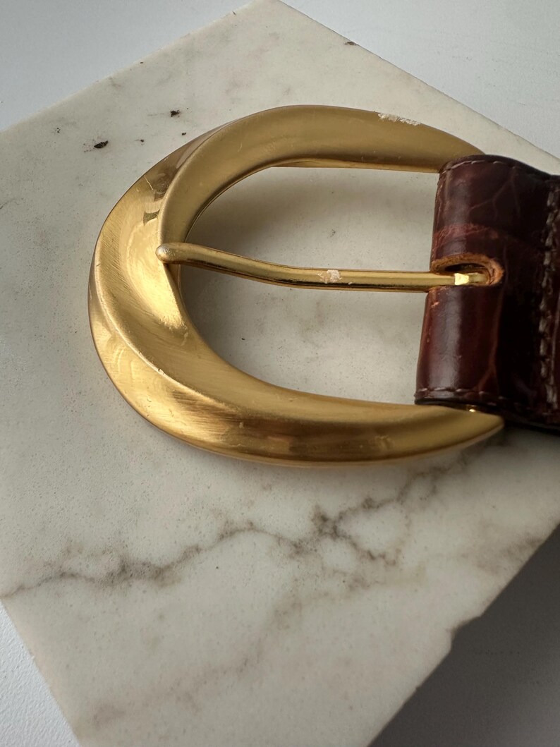 Puede incluir: Primer plano de un cintur&oacute;n de cuero marr&oacute;n con una hebilla grande de color dorado. La hebilla tiene forma de media luna curvada y un dise&ntilde;o simple y elegante. El cintur&oacute;n de cuero parece tener textura.