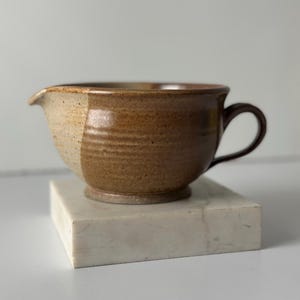 May include: A two-toned ceramic mixing bowl with a handle and a spout. The bowl is a combination of light tan and brown, with a dark brown handle. The bowl is sitting on a white square block.