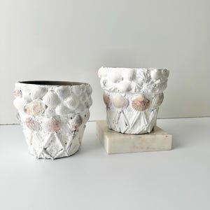 Może przedstawiać: Dwa białe, teksturowane doniczki ozdobione muszlami. Jedna doniczka stoi bezpośrednio na powierzchni, druga na małej, kwadratowej marmurowej podstawie. Doniczki mają rustykalną, nadmorską estetykę.