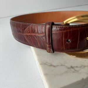 Puede incluir: Un cintur&oacute;n de cuero marr&oacute;n con una hebilla dorada. El cintur&oacute;n tiene un patr&oacute;n texturizado similar al de un cocodrilo. La hebilla tiene un dise&ntilde;o grande y curvado. El cintur&oacute;n est&aacute; sobre una superficie de m&aacute;rmol blanco.