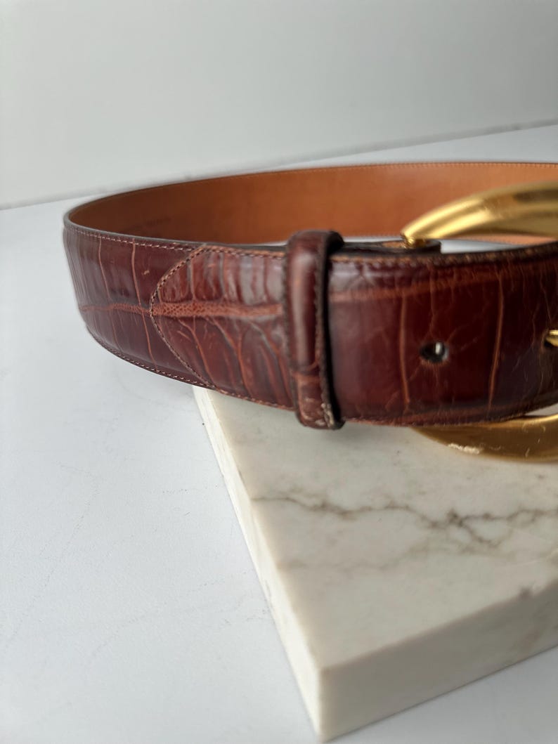 Puede incluir: Un cintur&oacute;n de cuero marr&oacute;n con una hebilla dorada. El cintur&oacute;n tiene un patr&oacute;n texturizado similar al de un cocodrilo. La hebilla tiene un dise&ntilde;o grande y curvado. El cintur&oacute;n est&aacute; sobre una superficie de m&aacute;rmol blanco.