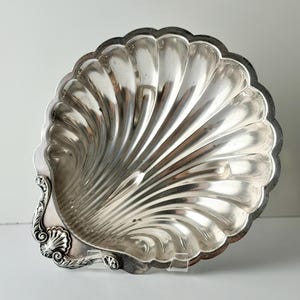 May include: A silver-toned metal dish shaped like a scallop shell. The interior features deep, radiating fluted grooves that extend from the base to the scalloped edge. The handle area is decorated with an ornate, embossed shell-like motif.