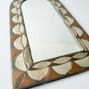 May include: An arched wall mirror featuring a decorative metal frame with alternating copper-toned triangular segments and silver-toned leaf-shaped pieces. The silver segments are embossed with a repeating curved line pattern throughout the border.