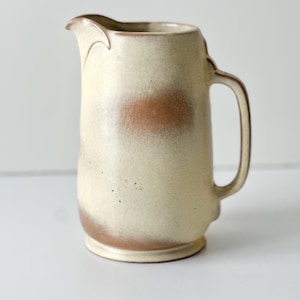May include: Cream-colored ceramic pitcher with a brown accent band at the base and a brown spot in the middle. The pitcher has a handle and a spout with a decorative edge. The handmade pottery has a rustic, speckled finish.
