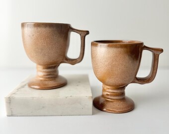 Pode incluir: Duas canecas de cerâmica marrom com base em pedestal. As canecas têm um esmalte salpicado e estão sobre uma superfície de mármore branco.