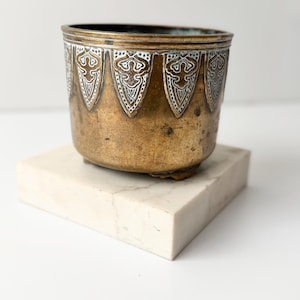 May include: A vintage brass planter with a decorative white and silver pattern around the rim. The planter sits on a white marble block, showcasing its aged patina and intricate design. A unique piece for home decor.