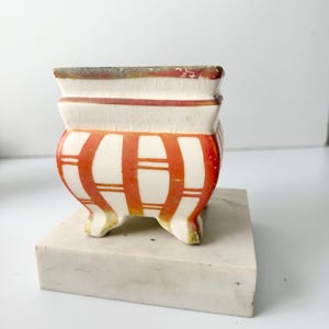 May include: A small, square ceramic planter with a white base and a crackled glaze finish. The exterior features vertical and horizontal orange stripes. The vessel is supported by four short, curved feet and rests on a rectangular marble block.