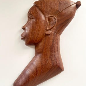 May include: A wooden sculpture of a person's head and neck, carved in a profile view. The sculpture is a dark brown color and has a smooth, polished finish.