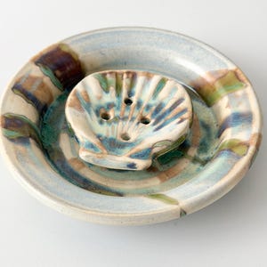 Può includere: Un portasapone rotondo in ceramica con un inserto rialzato a forma di conchiglia dotato di tre fori di drenaggio. Il pezzo è rifinito con uno smalto variegato nei toni del blu, verde e marrone, creando un effetto marmorizzato sulla superficie.