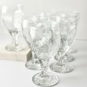 May include: A set of clear, textured water goblets. The glasses feature a distinctive hammered glass design on the bowl, stem, and base. The goblets are arranged on a white surface, with one goblet slightly separate from the others.