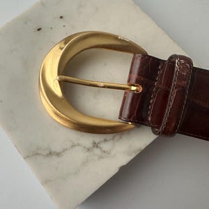 Puede incluir: Un cintur&oacute;n de cuero marr&oacute;n con una hebilla dorada. La hebilla tiene un dise&ntilde;o grande y curvado con una sola espiga. El cintur&oacute;n tiene un patr&oacute;n texturizado similar al de un cocodrilo.