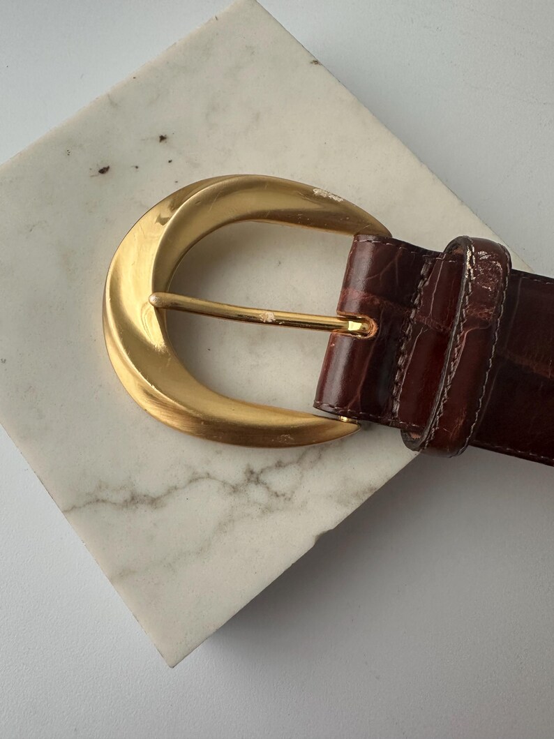 Puede incluir: Un cintur&oacute;n de cuero marr&oacute;n con una hebilla dorada. La hebilla tiene un dise&ntilde;o grande y curvado con una sola espiga. El cintur&oacute;n tiene un patr&oacute;n texturizado similar al de un cocodrilo.