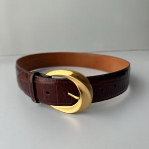 Puede incluir: Un cintur&oacute;n de cuero marr&oacute;n con una hebilla dorada. El cintur&oacute;n tiene un patr&oacute;n texturizado, similar al de un cocodrilo. La hebilla tiene una forma grande y redondeada con una sola espiga. El cintur&oacute;n est&aacute; dise&ntilde;ado para usar en la cintura.