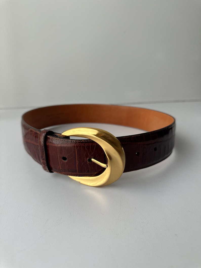 Puede incluir: Un cintur&oacute;n de cuero marr&oacute;n con una hebilla dorada. El cintur&oacute;n tiene un patr&oacute;n texturizado, similar al de un cocodrilo. La hebilla tiene una forma grande y redondeada con una sola espiga. El cintur&oacute;n est&aacute; dise&ntilde;ado para usar en la cintura.