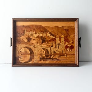 May include: Wooden serving tray with a marquetry landscape scene. The tray features a detailed depiction of a bridge, buildings, and mountains, all crafted from various wood tones. The tray has a dark brown frame and metal handles.