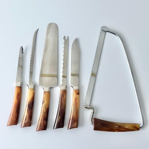 以下が含まれることがあります： ヴィンテージのサービング用具のコレクション。ナイフとノコギリのような道具が含まれています。刃はシルバー色で、ハンドルはマーブル模様の茶色とタン色です。白い表面に配置されています。