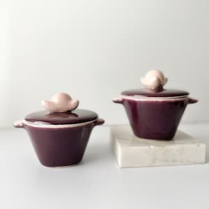 Può includere: Due piccoli vasetti in ceramica viola con coperchi rosa. I vasetti hanno una forma arrotondata e sono appoggiati su una superficie di marmo bianco.