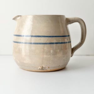 May include: A beige ceramic pitcher with a rounded body and a single handle. The exterior features two horizontal blue stripes encircling the middle section. The surface has a glossy finish with a small chip visible near the base of the vessel.