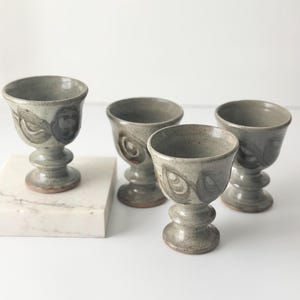 Puede incluir: Cuatro copas de cerámica hechas a mano en un tono gris verdoso apagado. Cada copa presenta una base de pedestal y una parte superior en forma de cuenco con un patrón de remolino decorativo. Las copas están dispuestas sobre una superficie blanca.