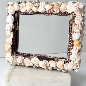 Op de afbeelding: Rechthoekige spiegel met een lijst versierd met diverse schelpen in wit, beige en bruin. De lijst staat op een witte marmeren voet, wat een maritieme sfeer creëert. De spiegel reflecteert de omgeving.
